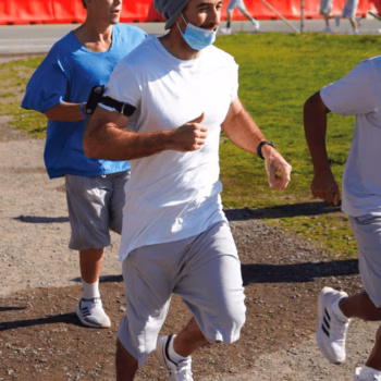 A man in a white t-shirt, grey shorts, and a blue beanie runs on a dirt path, wearing a light blue face mask and an armband. Other runners are visible in the background.