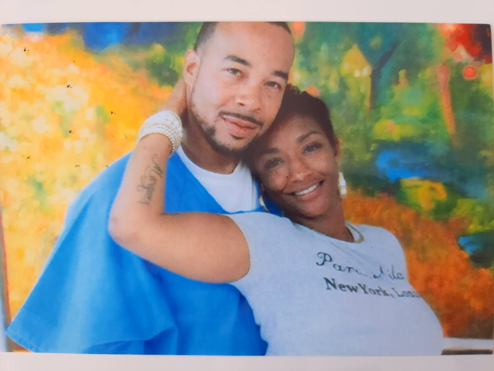 A man and a woman posing together in front of a colorful painting. The woman wears a light blue t-shirt with 'Paris, New York, London' printed on it.