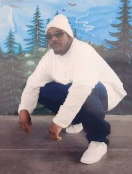 Michael 2 crouches in a white outfit and sunglasses against a painted forest backdrop.