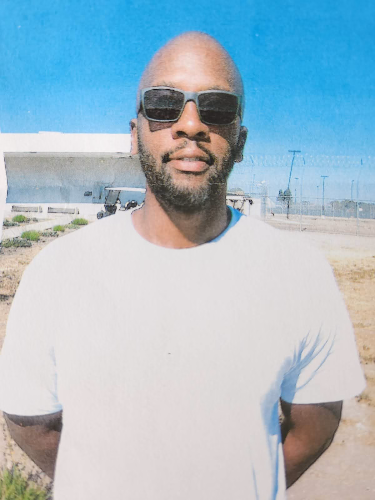 Reinald, a man with a beard and sunglasses, stands in front of a modern building under a clear blue sky.