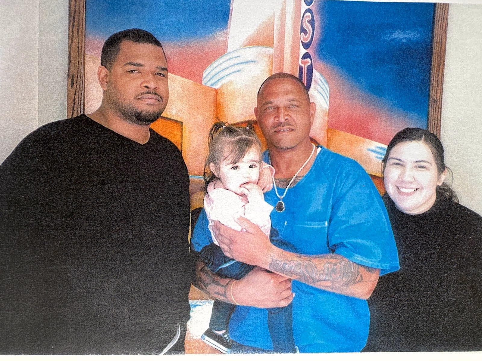 Four people, two men, a woman, and a baby, pose together in front of a colorful mural.