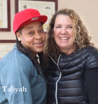 Taliyah, wearing a red cap and blue shirt, stands smiling next to another woman in a black vest, with framed accreditation certificates on the wall behind them.