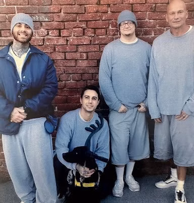 Men in prison clothes with a dog Four men in grey prison-style clothing and one man kneeling with a black dog, all posing in front of a red brick wall.