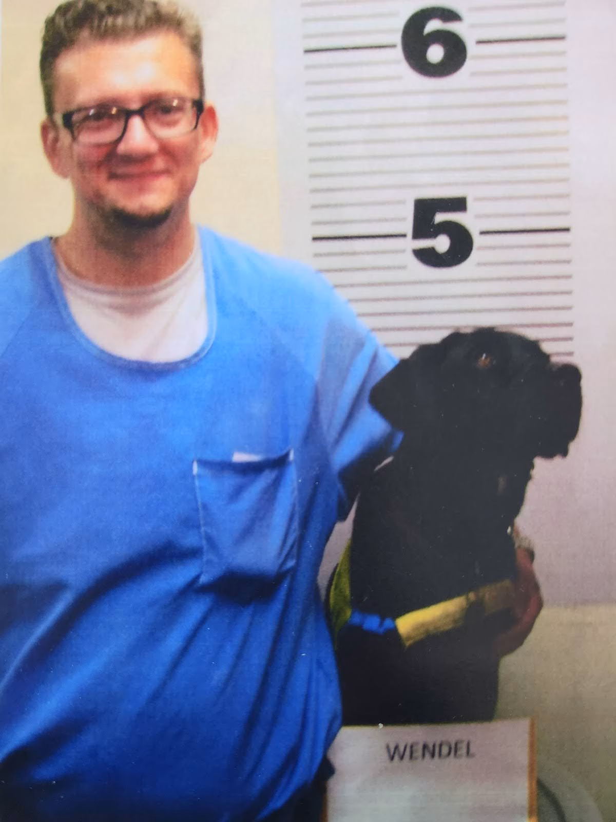 A man in a blue shirt stands next to a black dog named Wendel in front of a height chart.