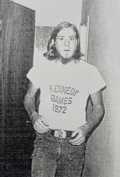 unnamed-2 A man with long hair and a beard wearing a white t-shirt that reads 'KENNEDY GAMES 1972' and jeans.