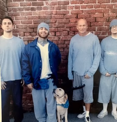 unnamed Four men in light blue prison uniforms stand against a brick wall with a small dog.
