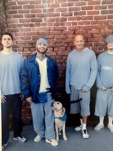 unnamed Four men in light blue prison uniforms stand against a brick wall with a small dog.