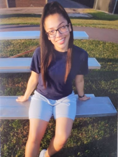 A young woman with long dark hair and glasses smiles while sitting on a concrete bench outdoors.
