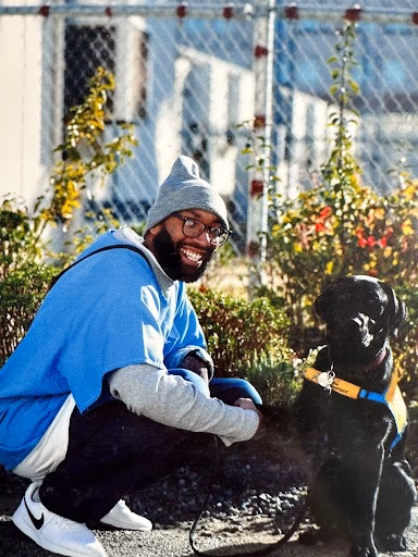 A smiling man in a blue hoodie and grey beanie crouches next to a black dog wearing a yellow harness in a garden.