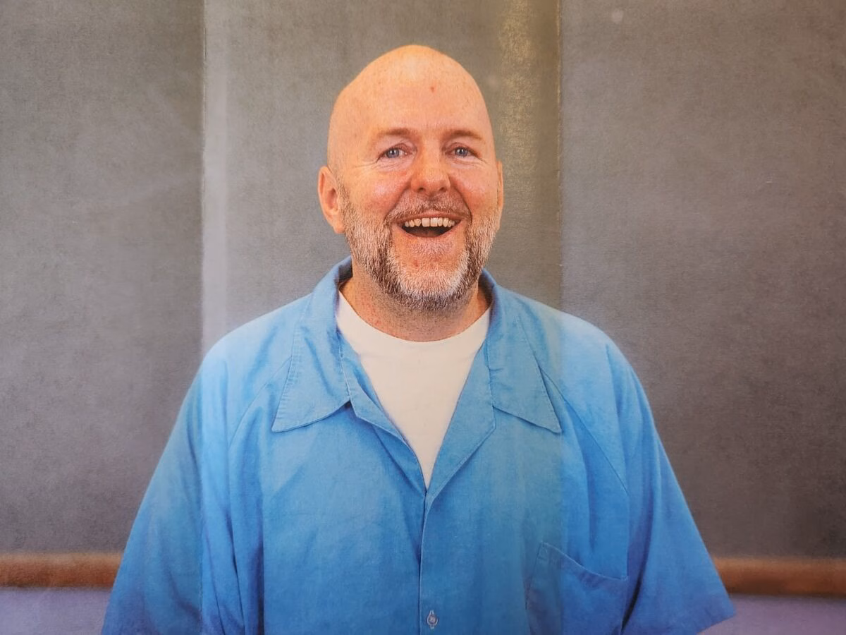 Portrait of James A bald man with a grey beard, wearing a blue shirt over a white t-shirt, smiles broadly at the camera against a grey wall.
