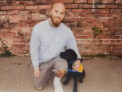 A bald man with a beard and tattoos kneels on a concrete surface next to a black dog wearing a blue and yellow vest, with a red brick wall in the background.