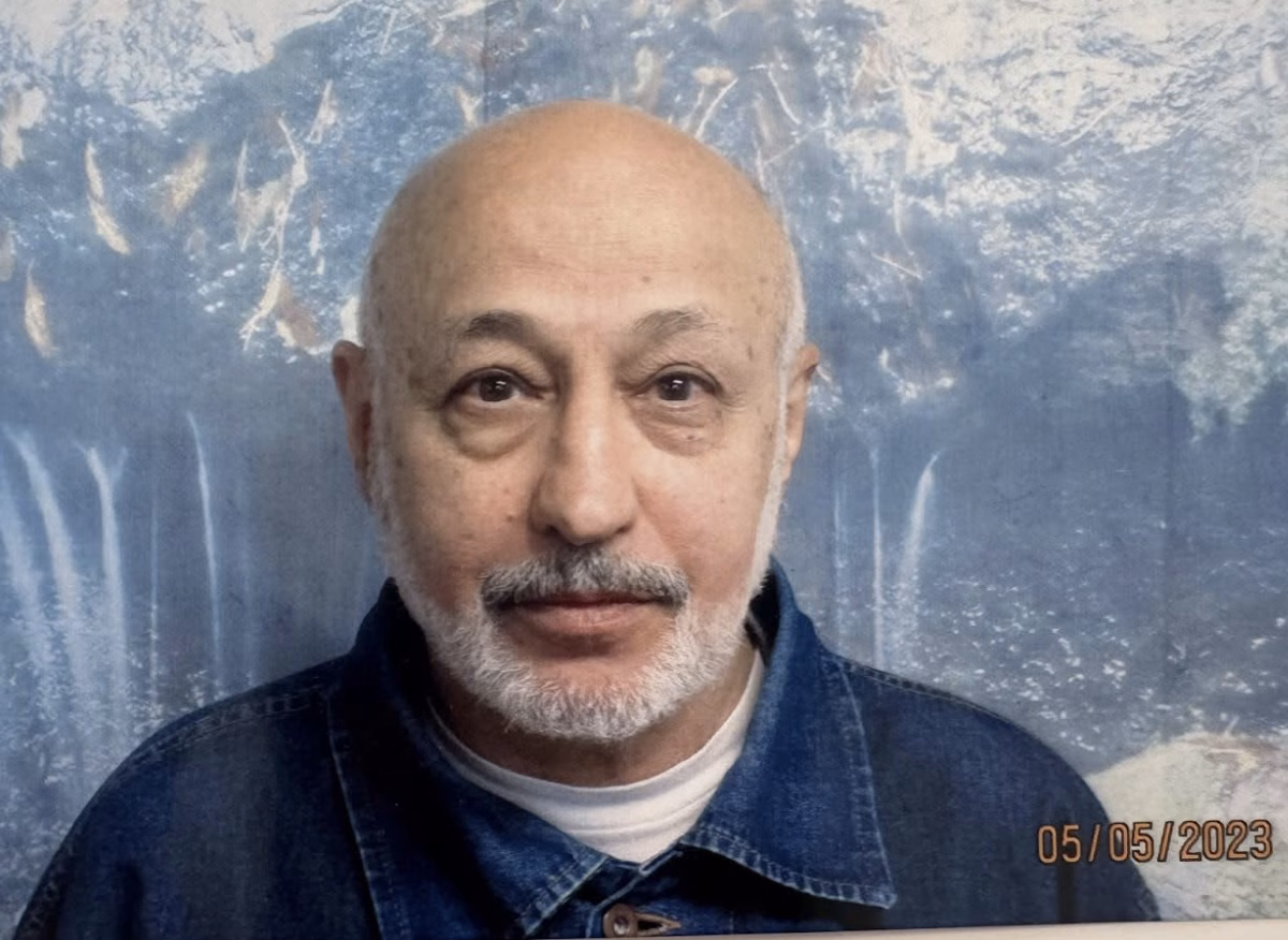A portrait of a bald man with a grey beard and mustache, wearing a blue denim jacket over a white shirt, against a blue textured background.