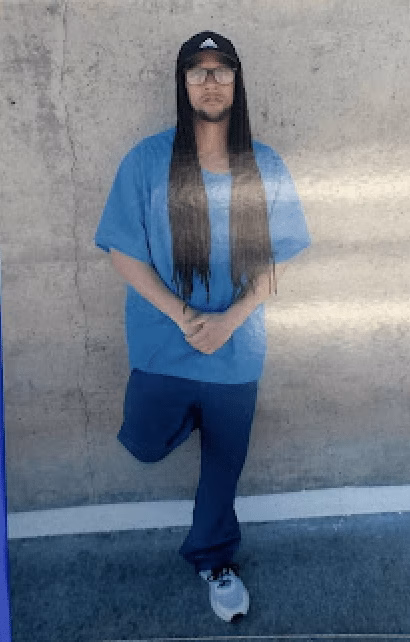 A person with long braids, glasses, and a cap stands against a concrete wall.