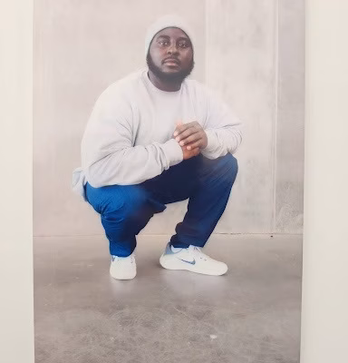 A man named Eric crouches against a plain wall, wearing a white beanie, sweatshirt, blue pants, and white Nike sneakers.
