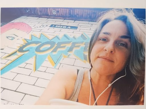 A woman with earphones takes a selfie in front of a large mural featuring the word 'COFFEE' in a comic book style.
