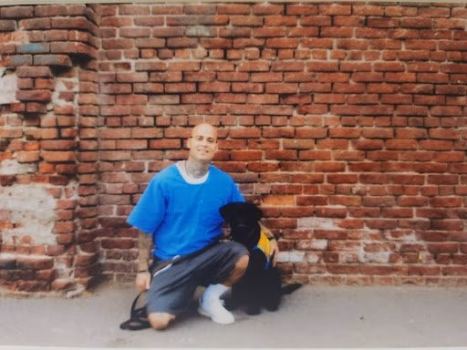 A man in a blue shirt and dark pants kneels on a concrete surface next to a black dog wearing a yellow vest, in front of a red brick wall.