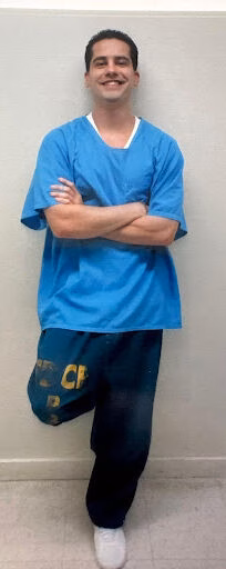 Randy stands with arms crossed, wearing blue scrubs and dark sweatpants.