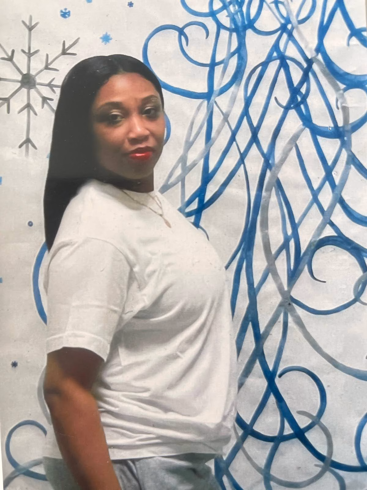 A woman with long dark hair in a white t-shirt stands in front of a wall decorated with blue swirls and snowflake patterns.