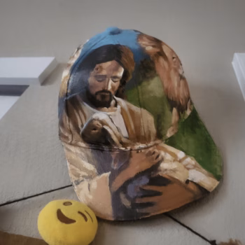 A baseball cap featuring a painted depiction of Jesus holding a child, resting on a surface next to a yellow smiley face ball.