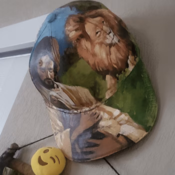 A baseball cap featuring a painted scene of Jesus and a lion, resting on a wooden surface next to a yellow smiley face ball.
