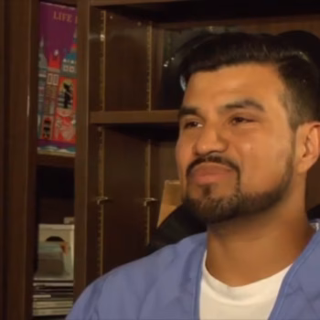 Miguel, a man with a beard in blue scrubs, is shown in a close-up shot against a wooden bookshelf filled with records and books.