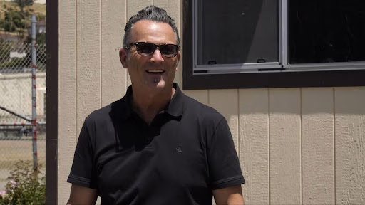 Brian King, wearing sunglasses and a black polo shirt, stands in front of a building.