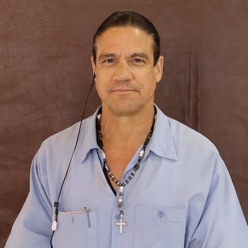 Bryan Mazza, a man with dark hair, wearing a light blue shirt, a beaded necklace with a cross, and an earpiece, against a brown background.