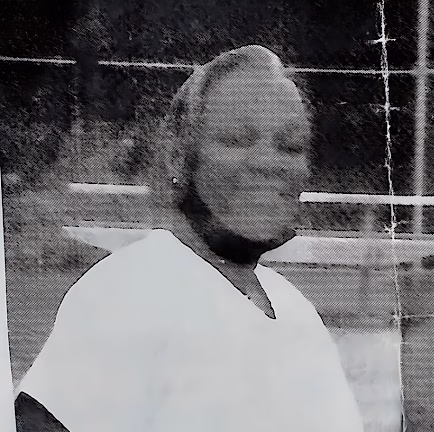 Black and white portrait of Mary P, a woman with short hair and a white top, standing in front of a fence.