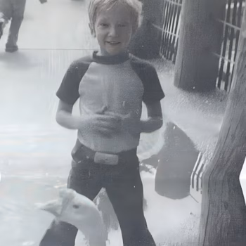 Shane Goddard 1 A young boy in a wetsuit stands in front of a glass enclosure, smiling as a dolphin swims by.