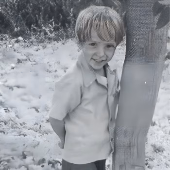 Shane Goddard 2 A young boy with short hair, wearing a light-colored shirt, smiles while leaning against a tree in a black and white outdoor setting.