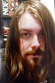 A close-up portrait of a man with long brown hair and a full beard, looking directly at the camera.