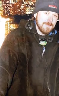 A man with a beard and a beanie stands in front of a city skyline at night.