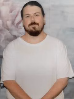 A man with a beard and dark hair wearing a white t-shirt, standing in front of a floral background.