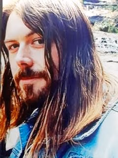 Close-up portrait of a man with long brown hair and a beard, wearing a blue denim jacket.