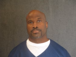 Toby Davis, a bald Black man with a goatee, wearing a blue V-neck shirt over a white undershirt, against a grey background.