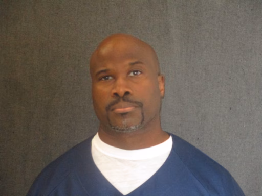 Toby Davis, a bald Black man with a goatee, wearing a blue V-neck shirt over a white undershirt, against a grey background.