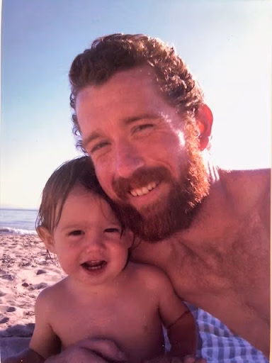 Travis George and a young child smiling together on a sandy beach under a bright sky.