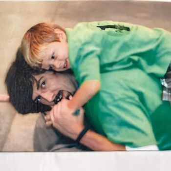 Lucas, 38, lying on his back while a young child in a green shirt playfully hugs his face.