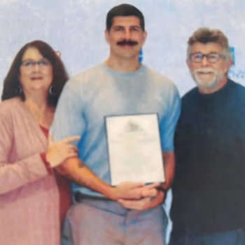 Lucas, 38, holding a framed certificate while standing between a woman and an older man.