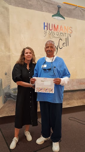 Ricky M, 64, in blue scrubs, receives a certificate from a woman in front of a 'Humans of San Quentin' mural.