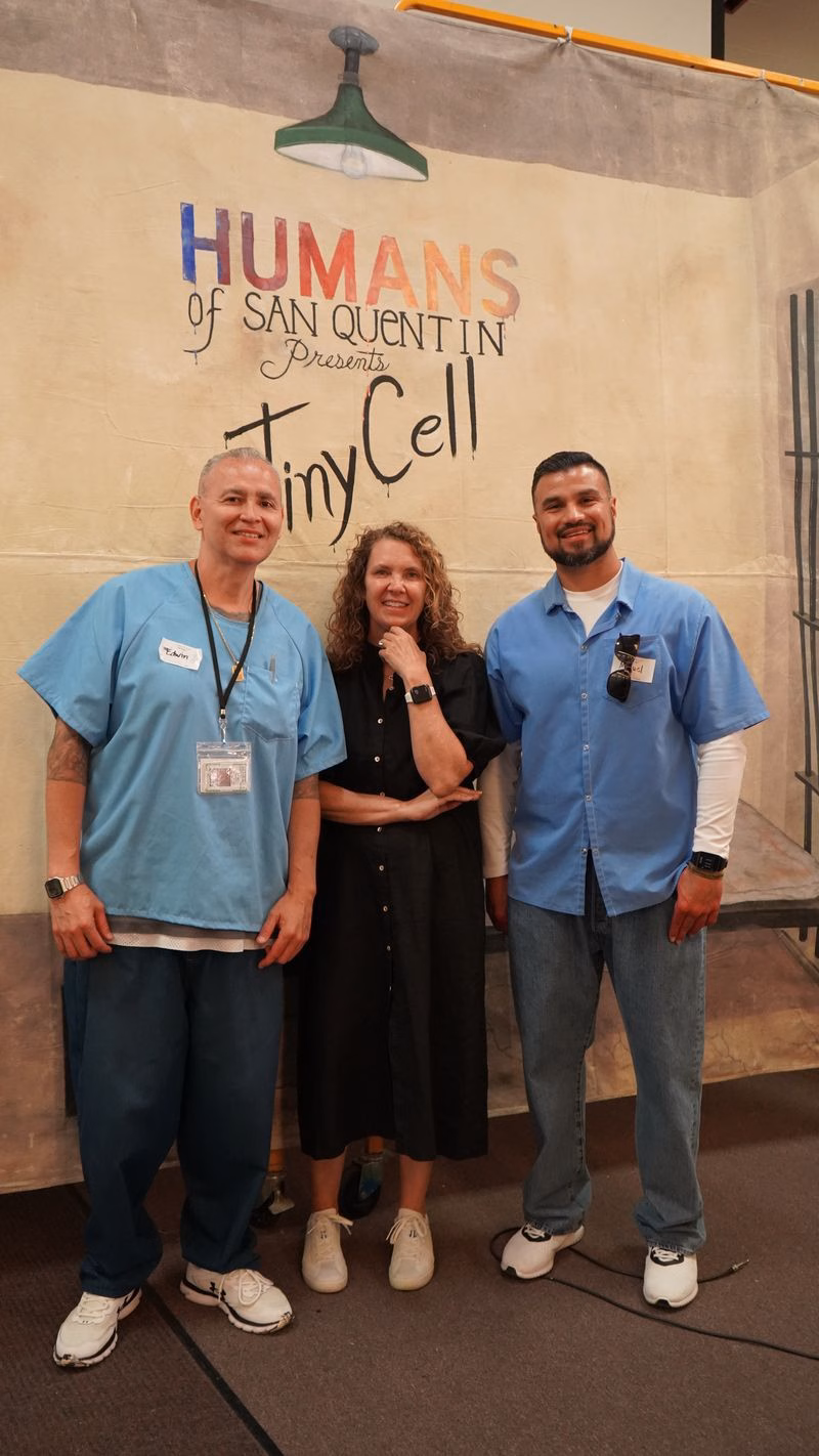 Three people in blue scrubs and a woman in a black dress stand in front of a banner for 'Humans of San Quentin Presents Tiny Cell'.
