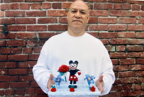 Francisco holds a white cake decorated with Mickey Mouse and Minnie Mouse figurines against a red brick wall.