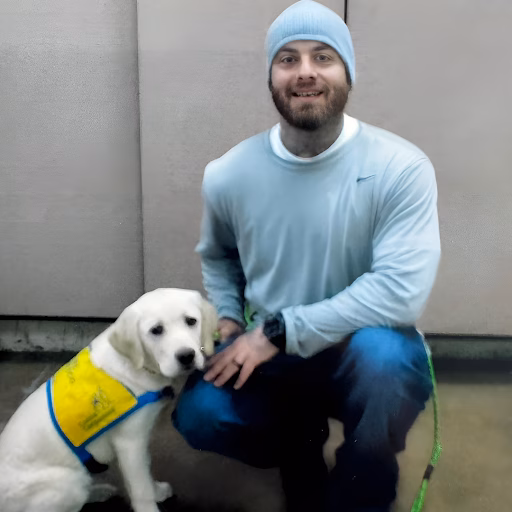 Jared kneeling and smiling next to a white Labrador puppy wearing a yellow service dog vest.