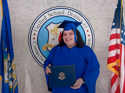 Katherine, a graduate in a blue cap and gown, smiles while holding a diploma in front of the Connecticut Unified School District #1 seal.