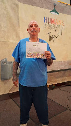 Michael Silva holds a certificate in front of a 'Humans of San Quentin' banner.
