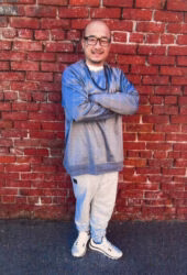 A bald man with glasses, Khiem, wearing a blue and grey sweater and light grey sweatpants, stands with arms crossed against a red brick wall.