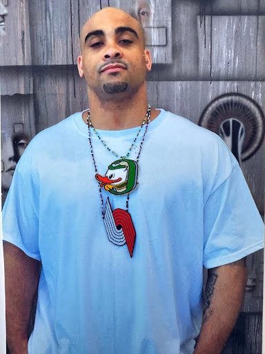 Antonio, 41, wearing a light blue t-shirt and a necklace with a cartoon duck pendant, stands against a wooden wall.