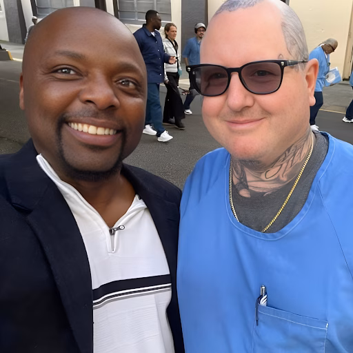 Coby, 50, a man with a shaved head and a dark blazer over a white shirt, stands next to a man with glasses and blue scrubs, both smiling outdoors.