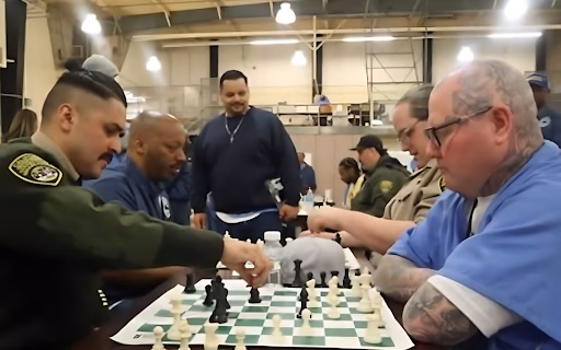 A man in a blue prison uniform plays chess with a uniformed officer in a large, industrial-style room.