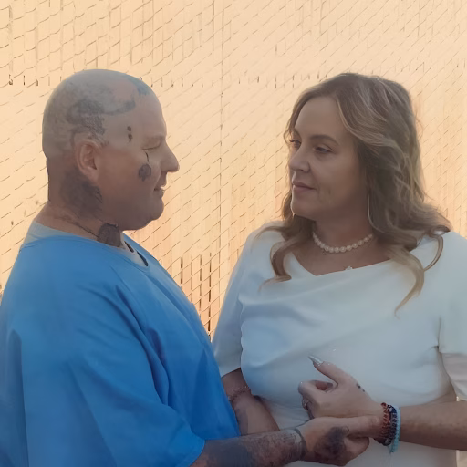 Coby, a man with extensive tattoos on his head and neck, stands in profile next to a woman in a white top, both looking at each other against a textured wall.
